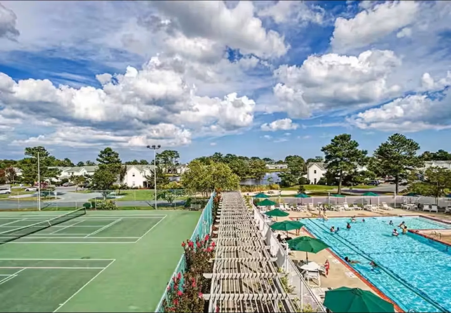 Elevated view of community amenities including outdoor tennis courts, a swimming pool with lounge area, and a pergola walkway under a cloudy blue sky.