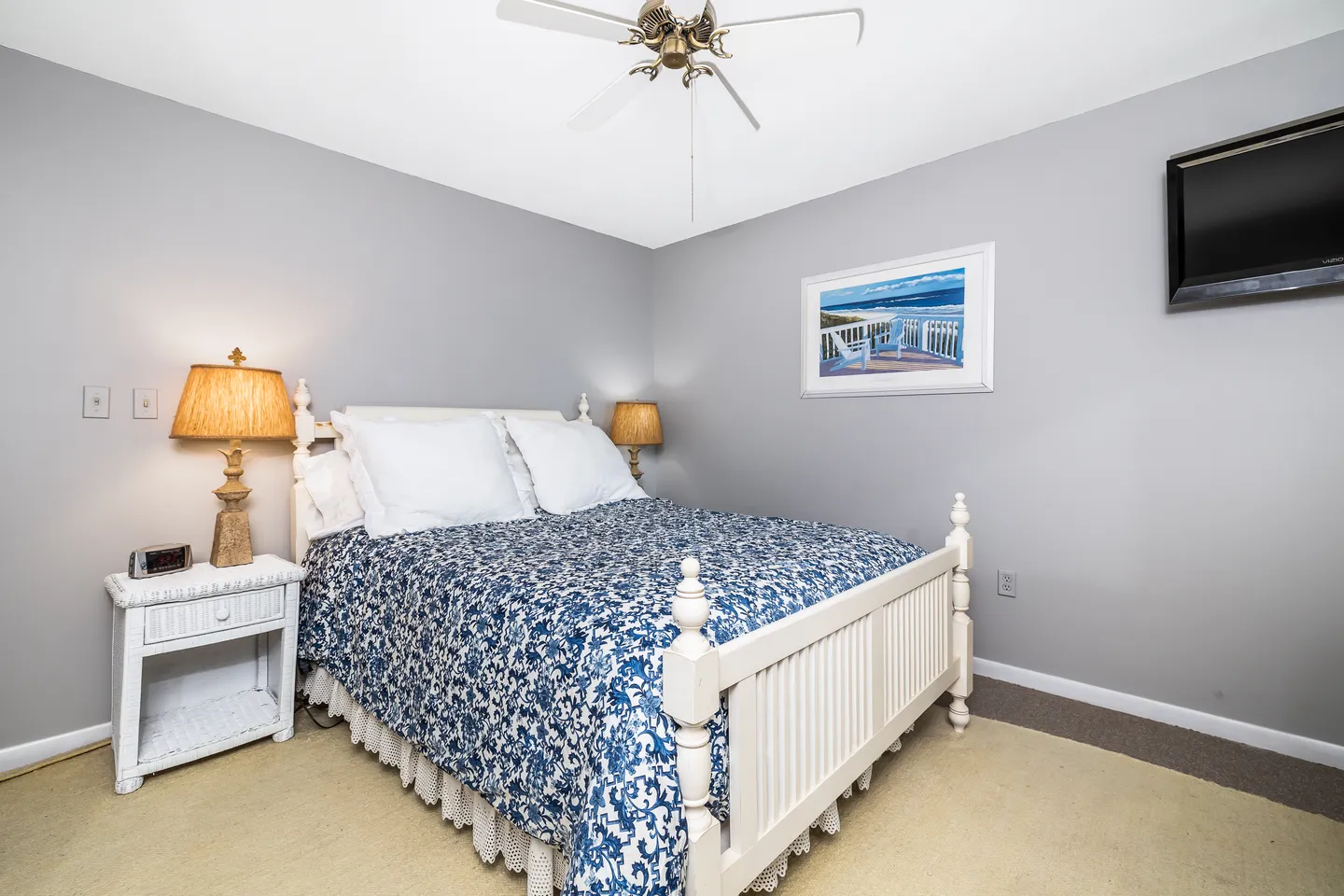 Bedroom with a white wooden bed frame, blue patterned quilt, white nightstand with lamp, and wall-mounted TV.