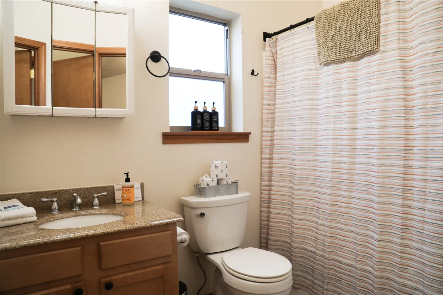 Bathroom with wooden vanity, speckled countertop, toilet, mirrored medicine cabinet, small window, and shower curtain.