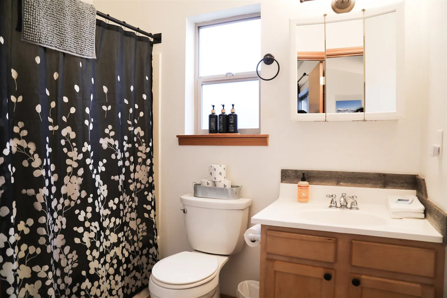 Bathroom showing a wooden vanity with white countertop, toilet, frosted window, and black patterned shower curtain.