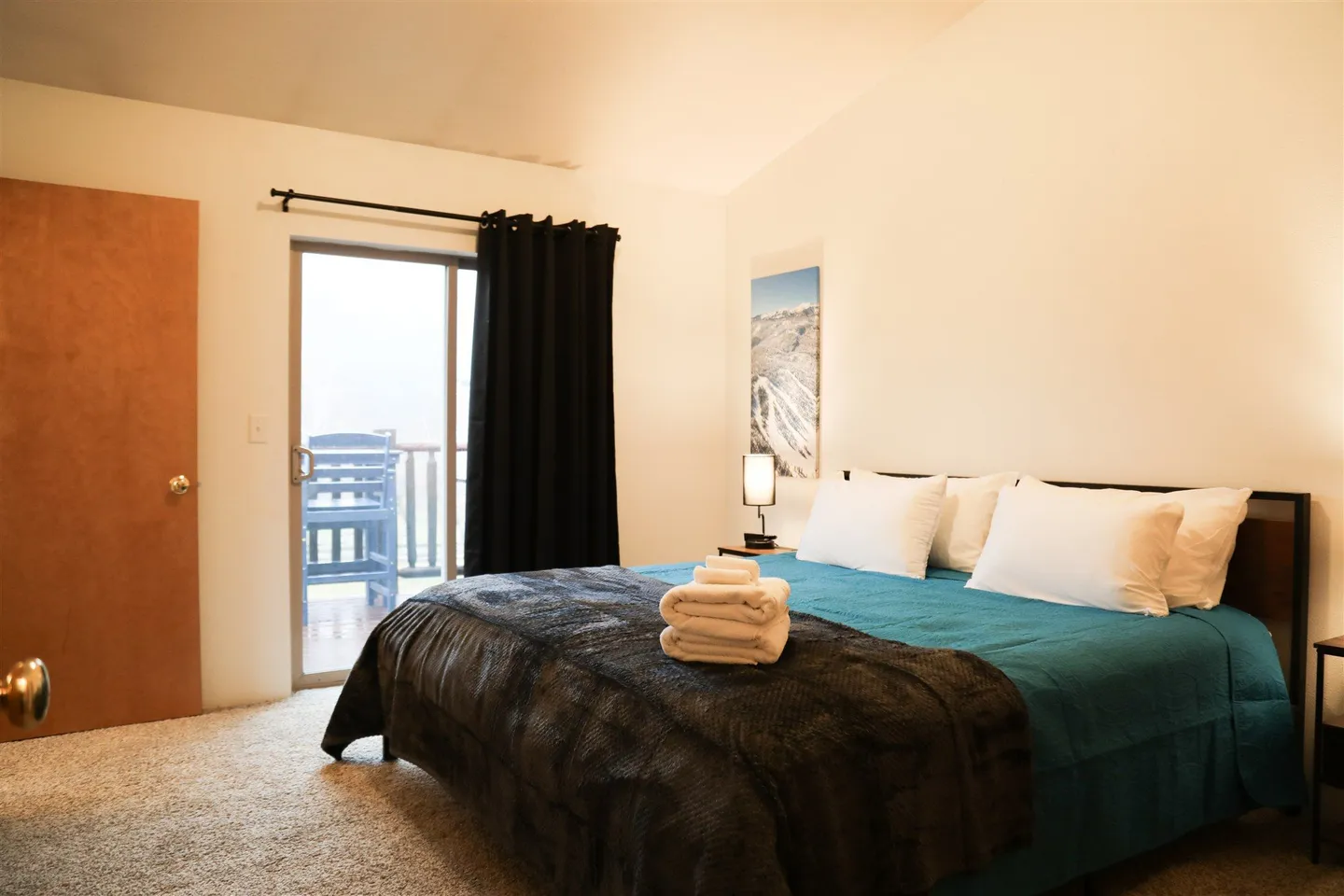 Bedroom featuring a made bed with teal and patterned blankets, carpeted flooring, and sliding glass doors opening to a balcony.