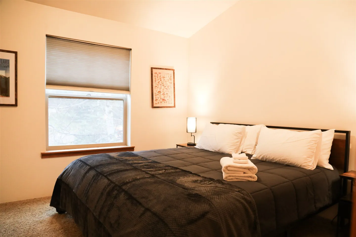 Bedroom featuring a made bed with dark bedding, white pillows, a stack of white towels, a window with blinds, and carpeted flooring.