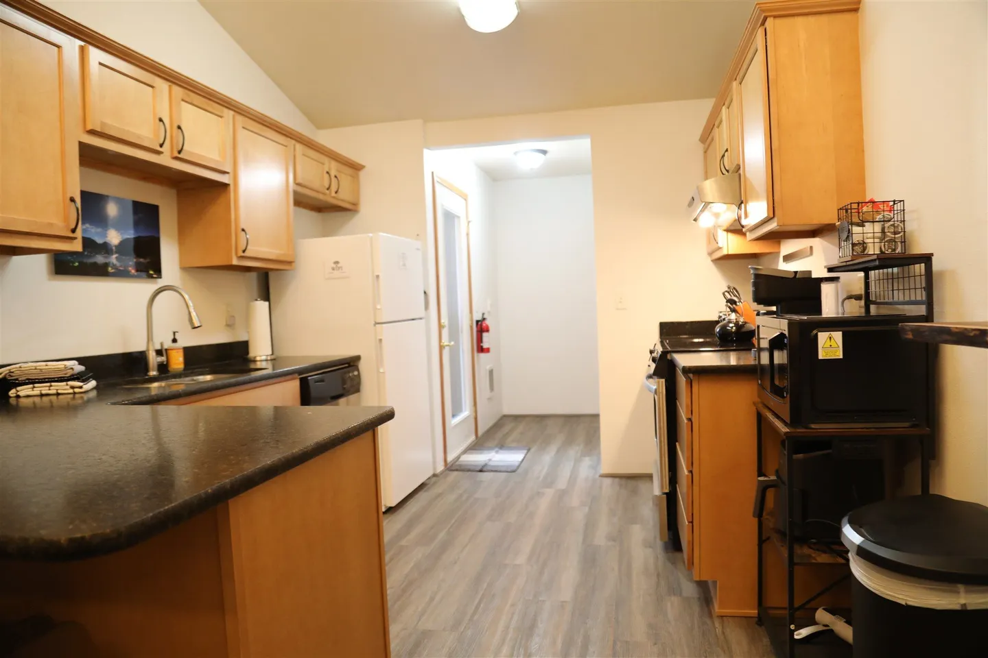 Kitchen with wooden cabinetry, dark countertops, white refrigerator, and view down a hallway with an exterior door.