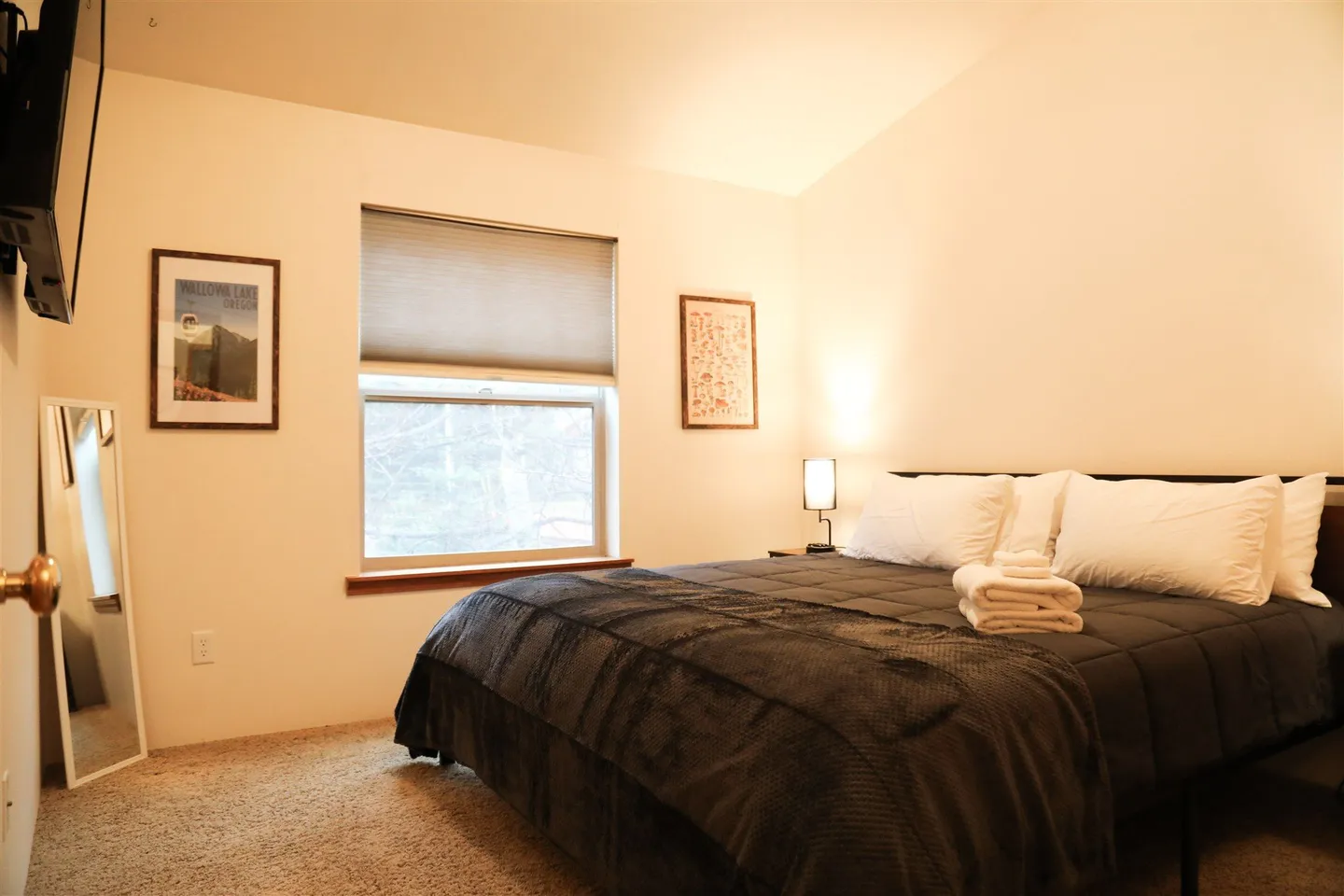 Bedroom with a bed made with a dark blanket, carpeted floor, window with blinds, and a full-length leaning mirror.