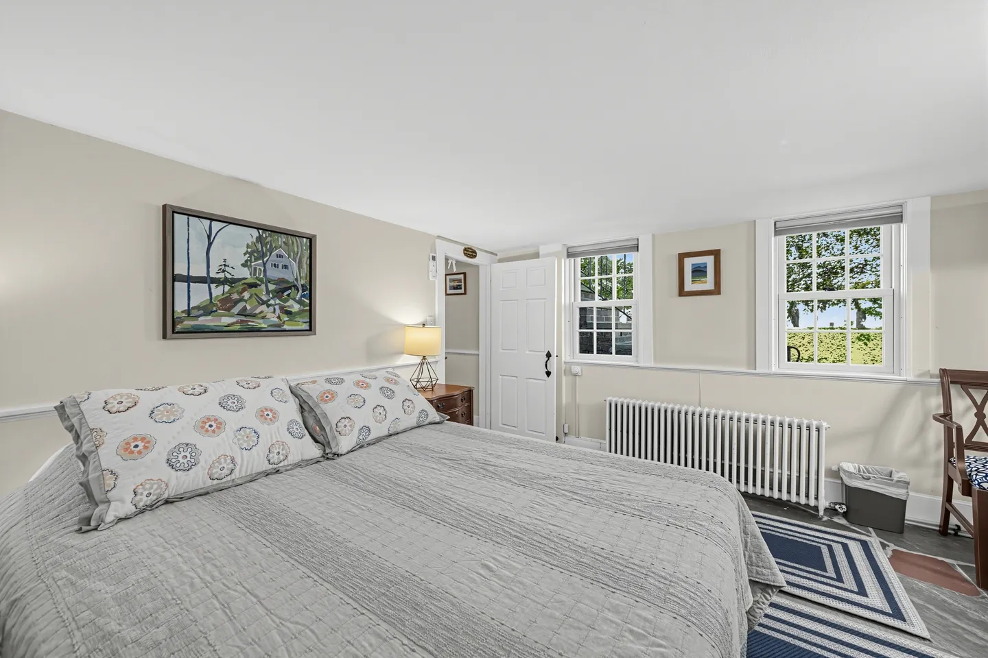 Bedroom with a large bed dressed in grey bedding, double-hung windows, a radiator, and framed art on the wall.
