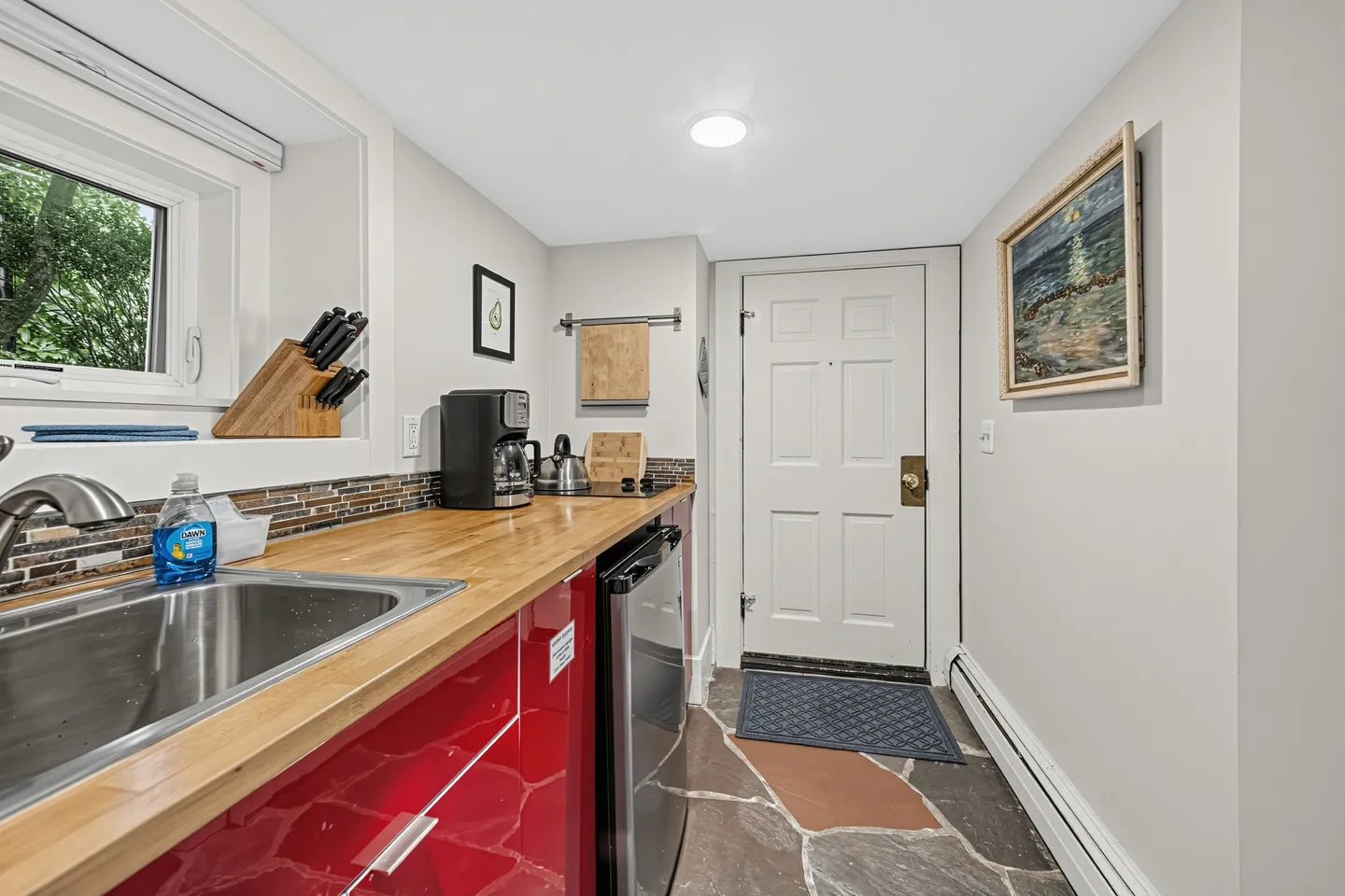 A kitchenette with glossy red cabinets, a wooden countertop, sink, mini fridge, and an exterior door with stone floor.