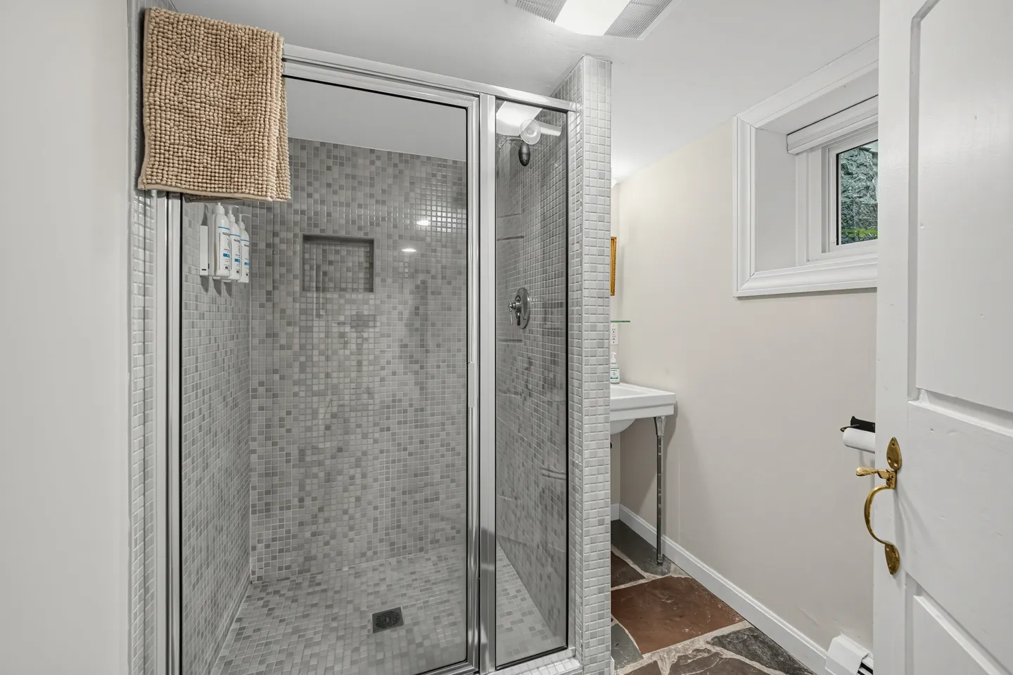 Bathroom with a glass-enclosed shower featuring small gray mosaic tiles next to a small white sink and stone floor.