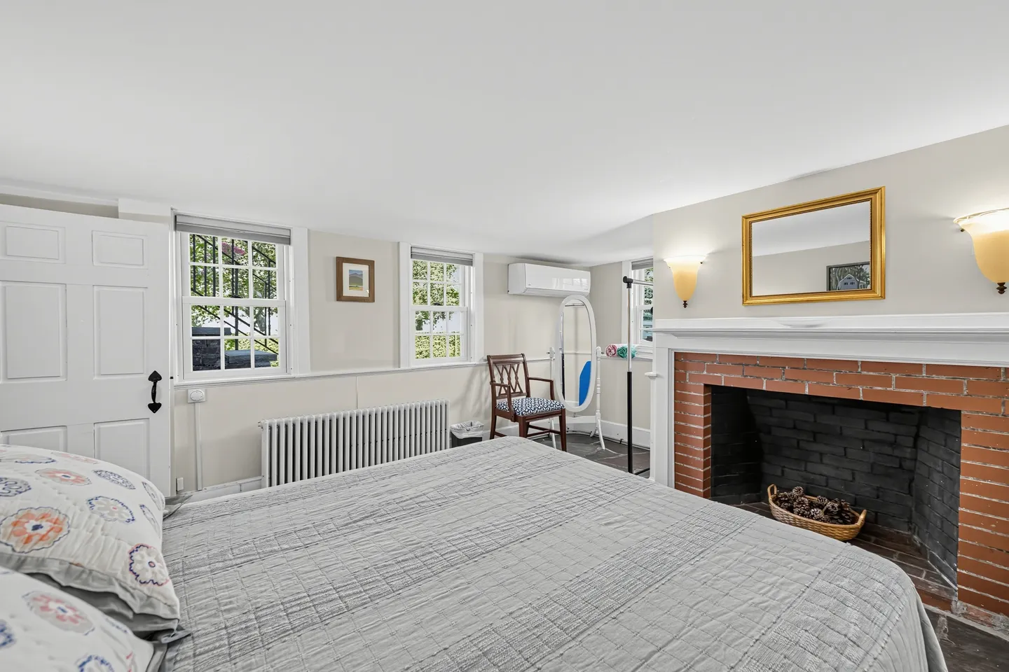 Bedroom with a bed in the foreground facing a brick fireplace, two windows, and an armchair.