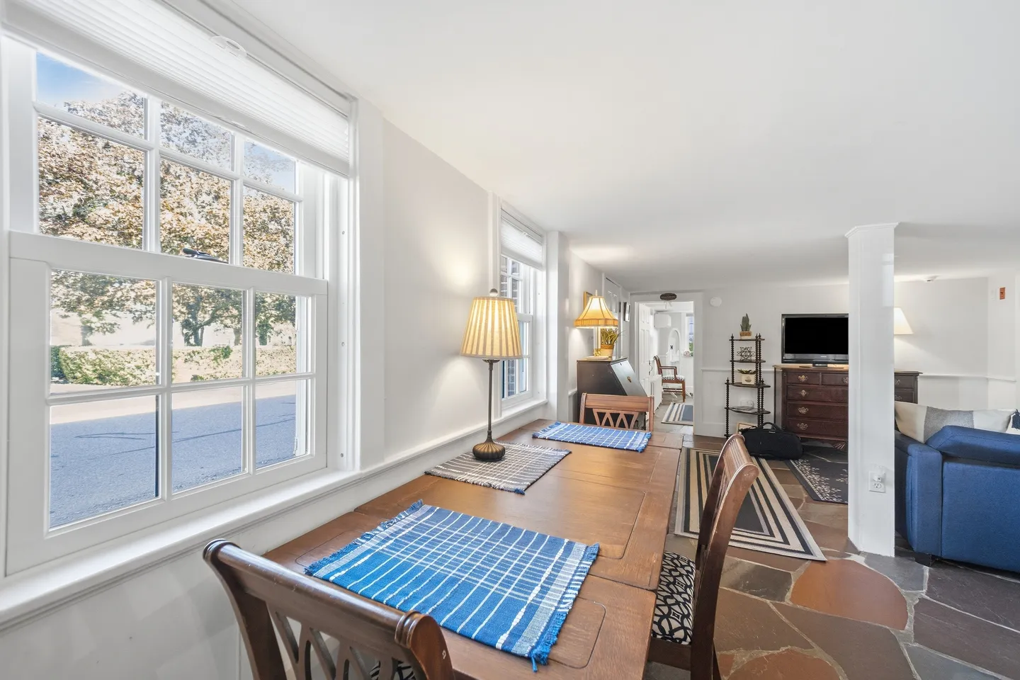 Dining table alongside large sash windows leading toward an open-concept living area with stone tile flooring.