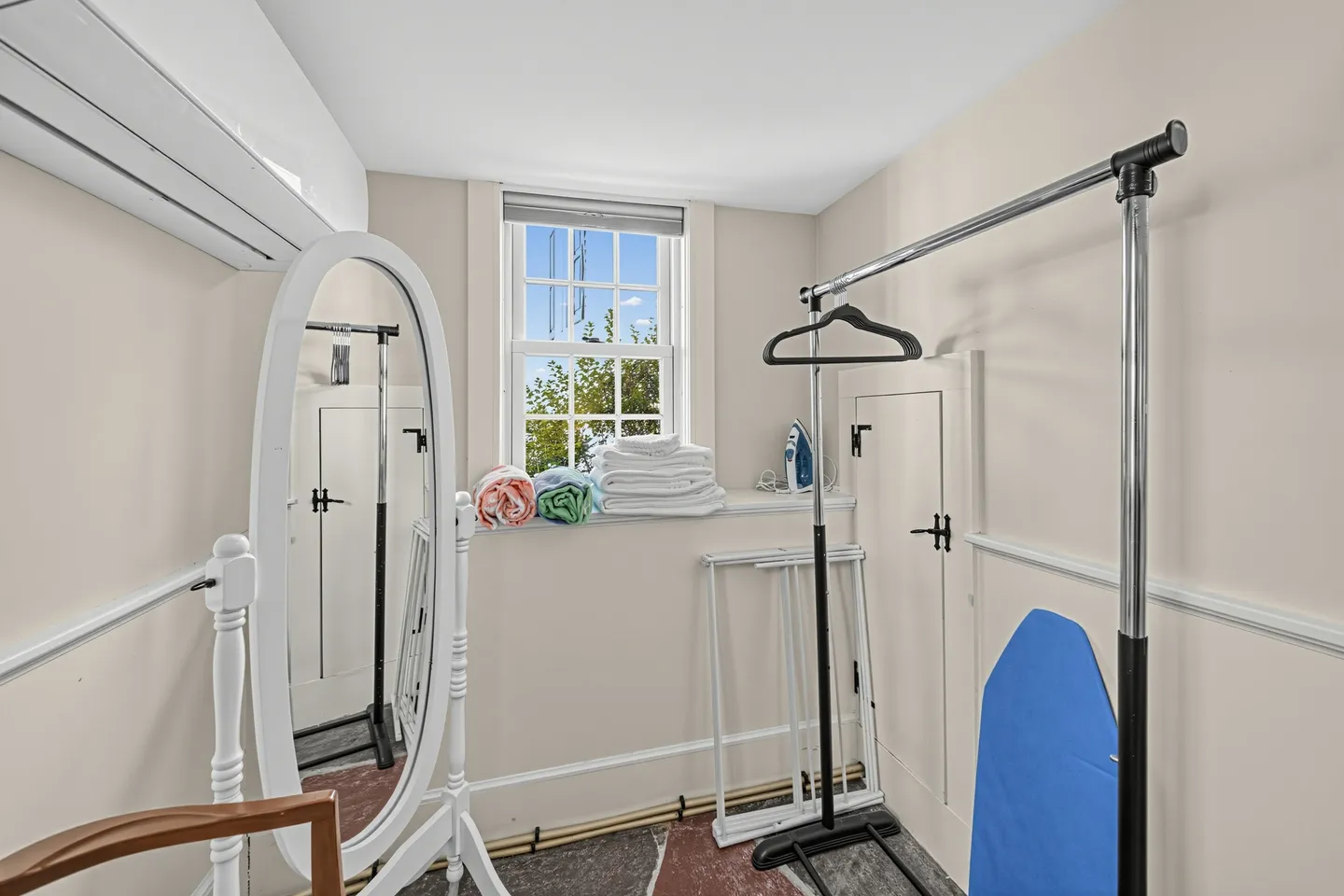 Dressing room or utility nook with a freestanding mirror, clothing rack, ironing board, and folded linens by a window.