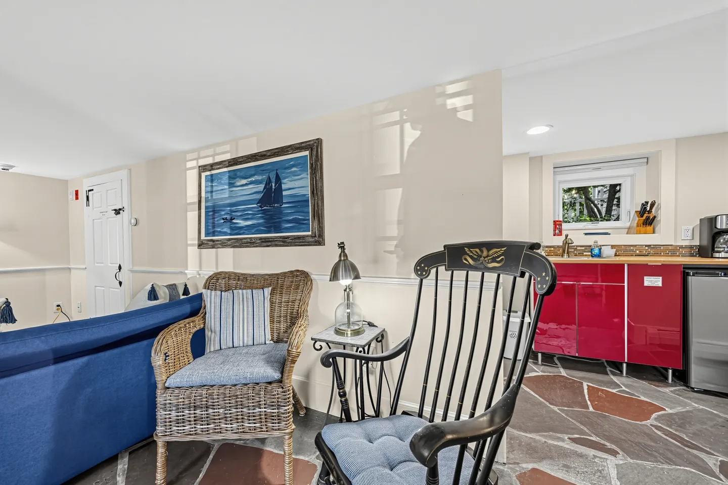 Seating area with a wicker chair and black rocking chair leading into a kitchenette with red cabinetry.