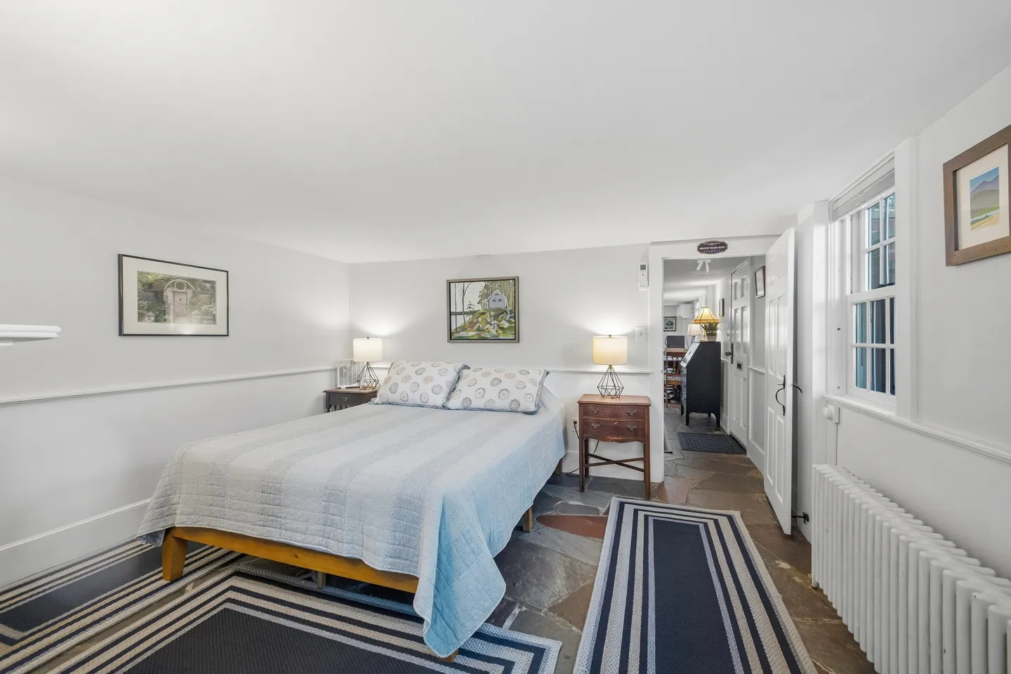 Bedroom with a bed, two nightstands with lamps, striped area rugs on a stone-look floor, and an open doorway leading down a hall.