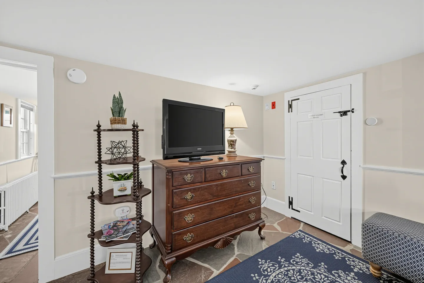 A corner of a room featuring a wooden dresser with a television, a corner shelf, a white door with black strap hinges, and stone-style flooring.