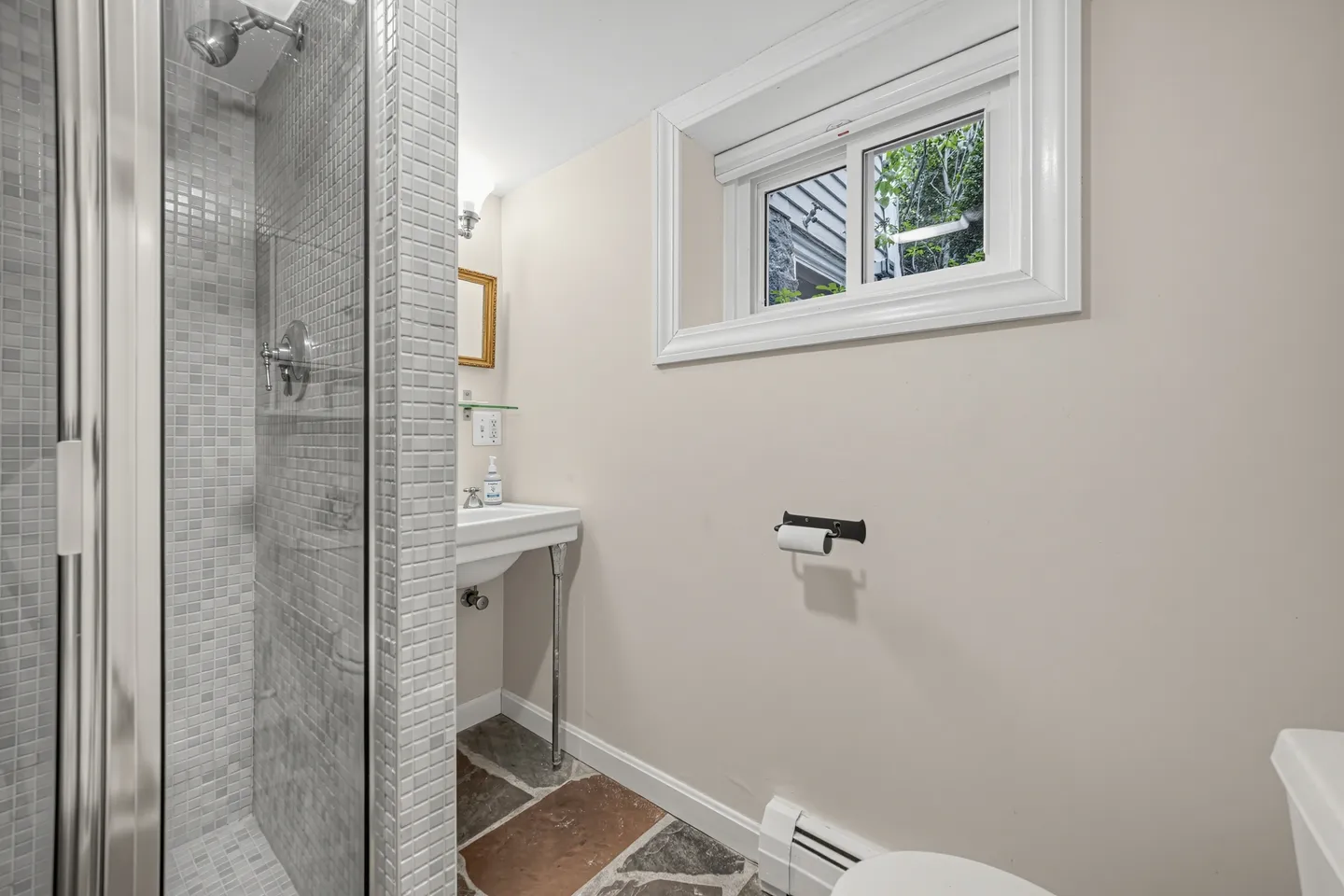 Bathroom showing a glass-enclosed mosaic tile walk-in shower, wall-hung sink, flagstone floor, and a small upper window.