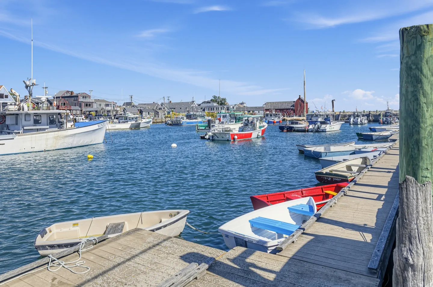 A harbor on a sunny day with small dinghies tied to a wooden dock and fishing boats anchored in calm water.