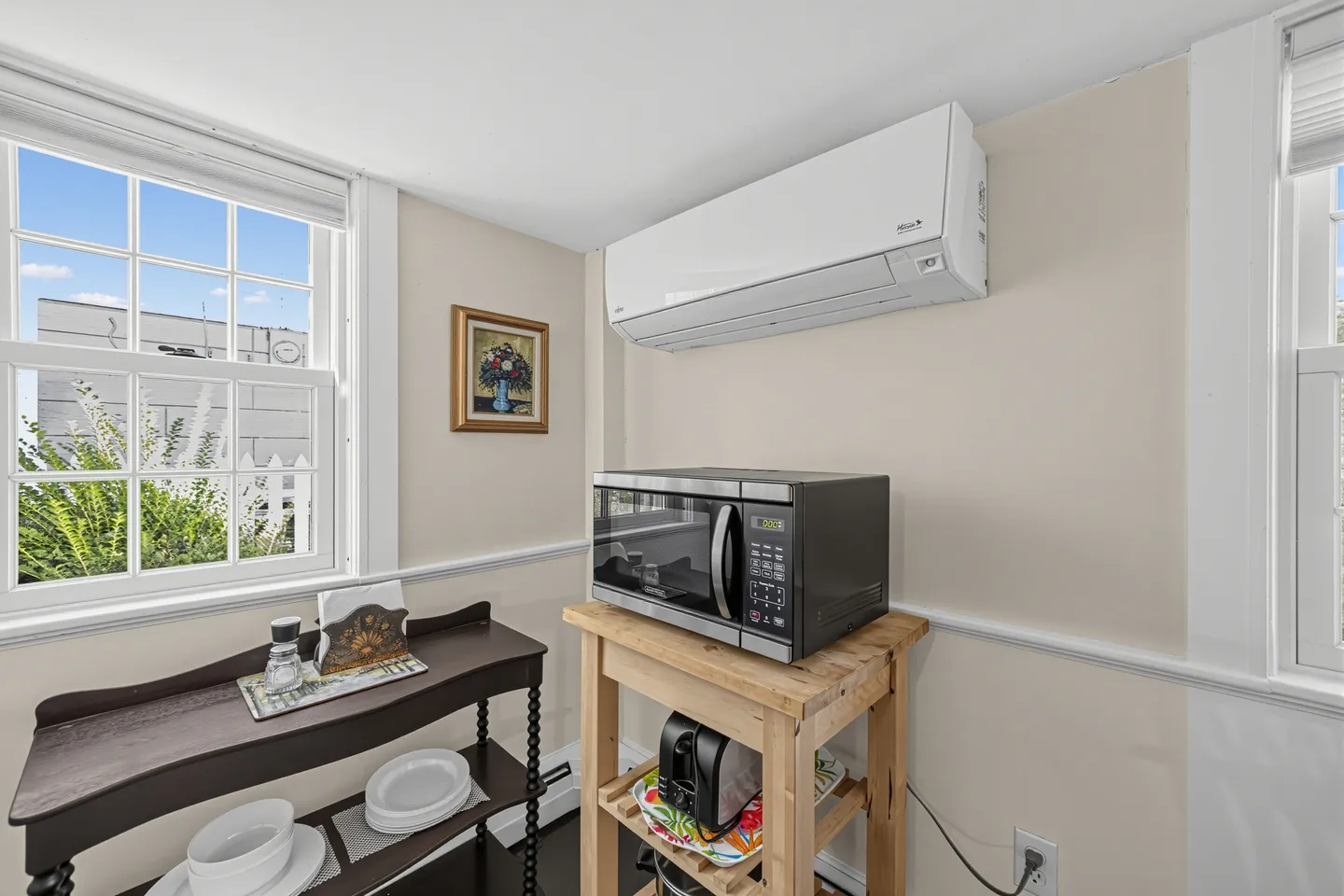 A corner of a room featuring a wooden cart with a microwave, a small side table holding dishes, and a wall-mounted ductless air conditioning unit.