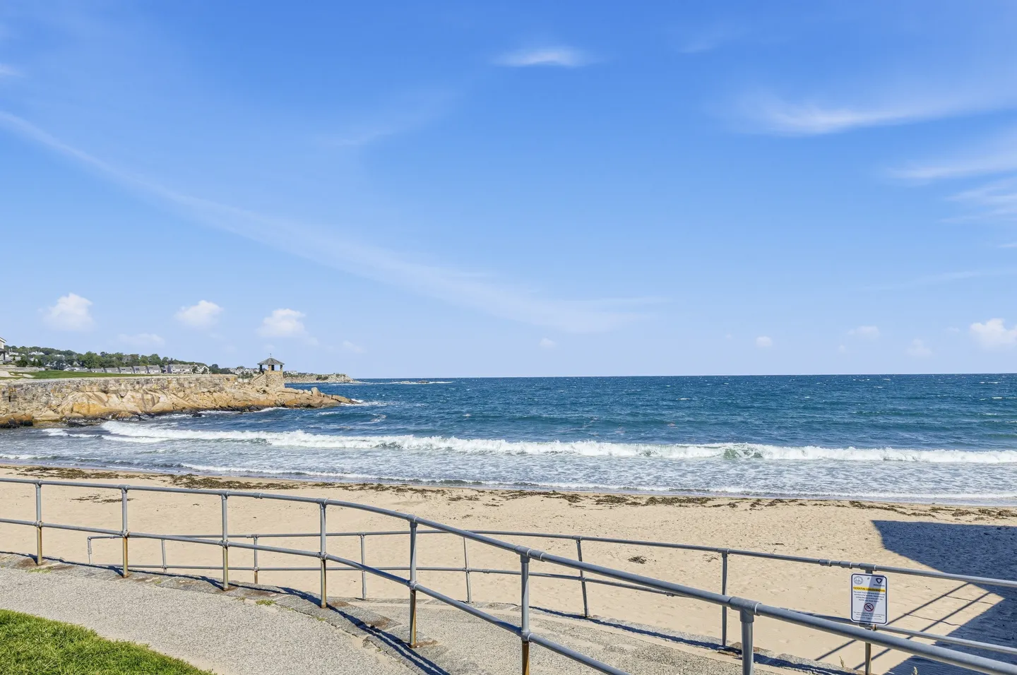 A sandy beach and blue ocean viewed from a paved path with metal railings under a clear sky.