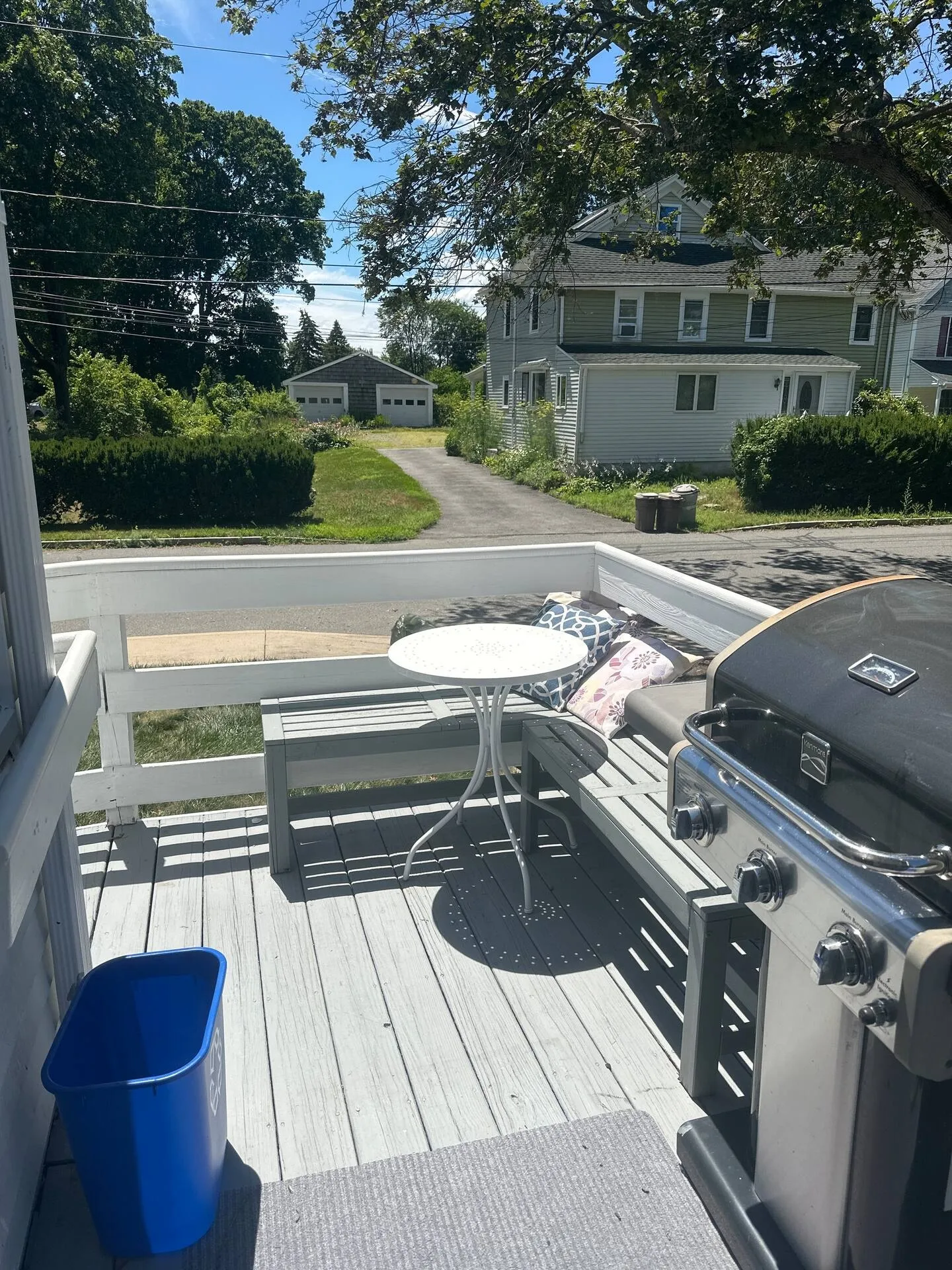 Outdoor deck with painted wood railings, bench seating, a round cafe table, and a gas grill overlooking a residential street.