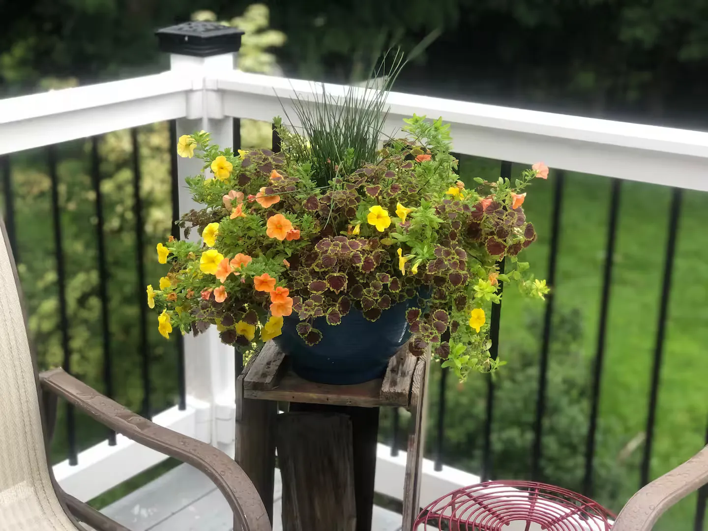 A potted planter filled with yellow and orange flowers and coleus sits on a rustic wooden stand on an outdoor deck.