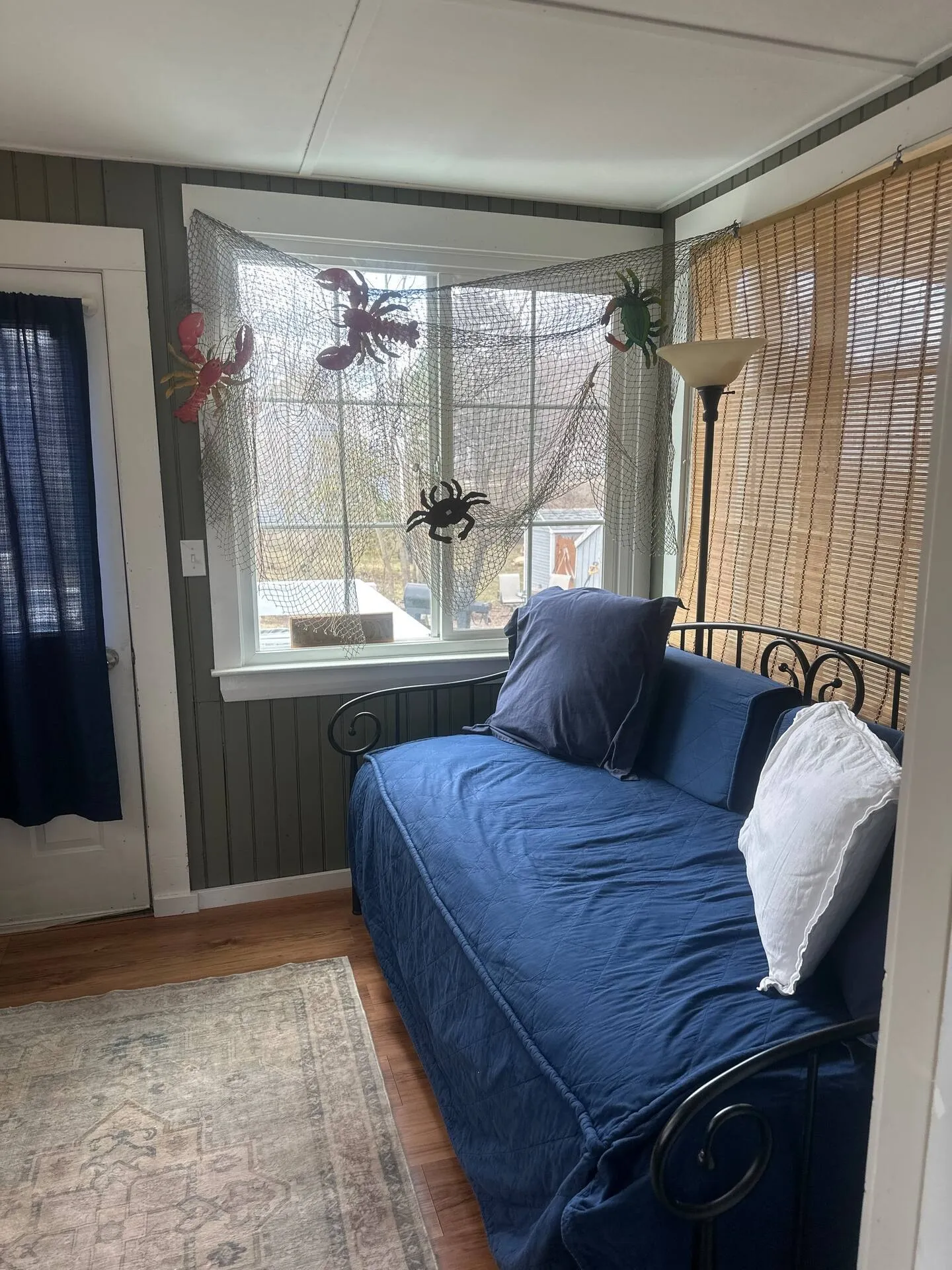 Sunroom corner with a wrought-iron daybed dressed in blue, wood paneled walls, and coastal decor over a window.