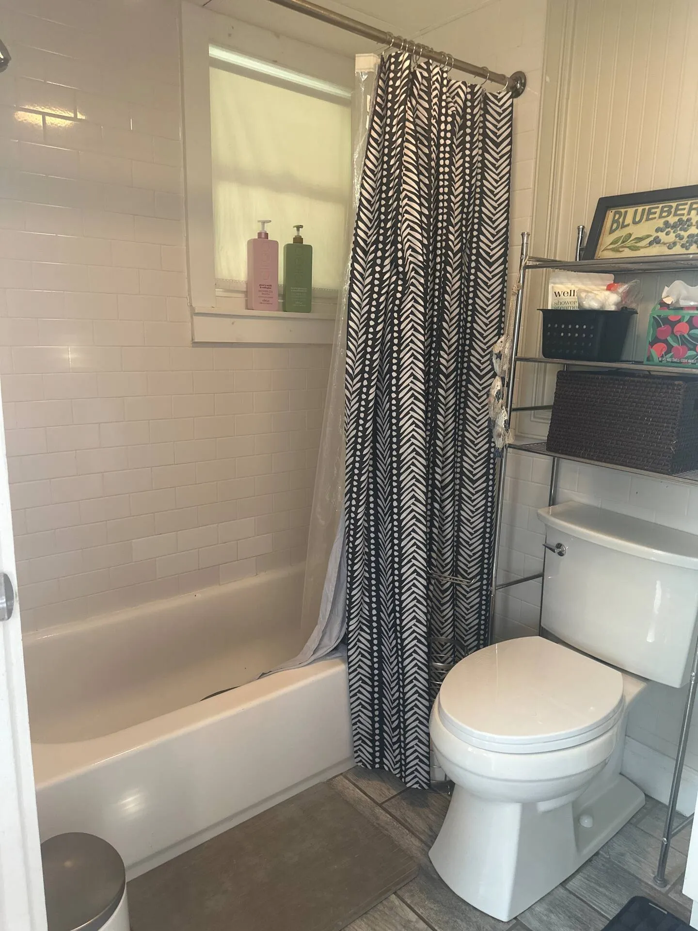 Bathroom showing a white bathtub with subway tile, a black and white patterned shower curtain, a window on the shower wall, and a toilet with an over-the-toilet metal shelf.