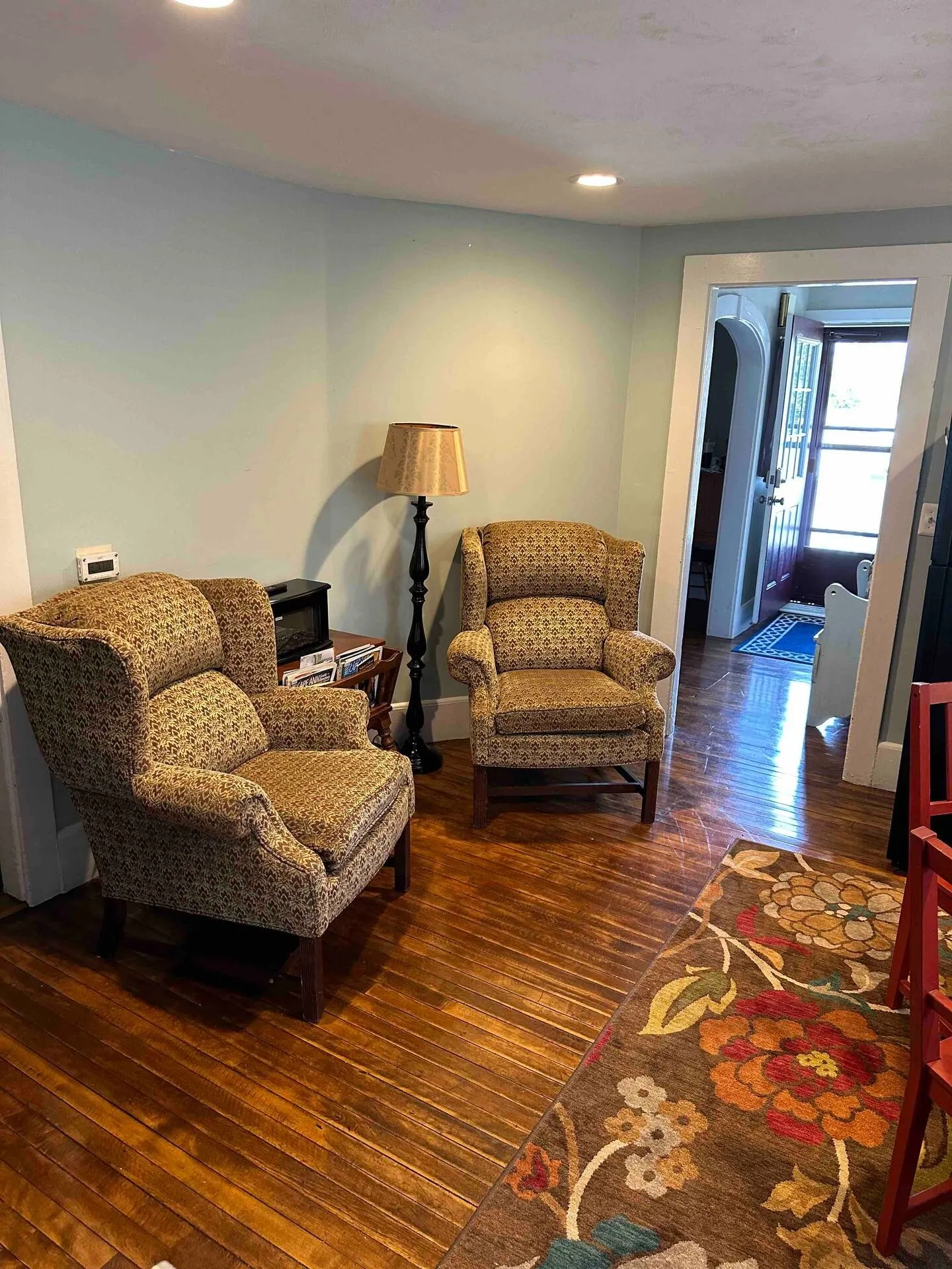Two patterned armchairs and a floor lamp in a room with hardwood floors leading toward an entryway.