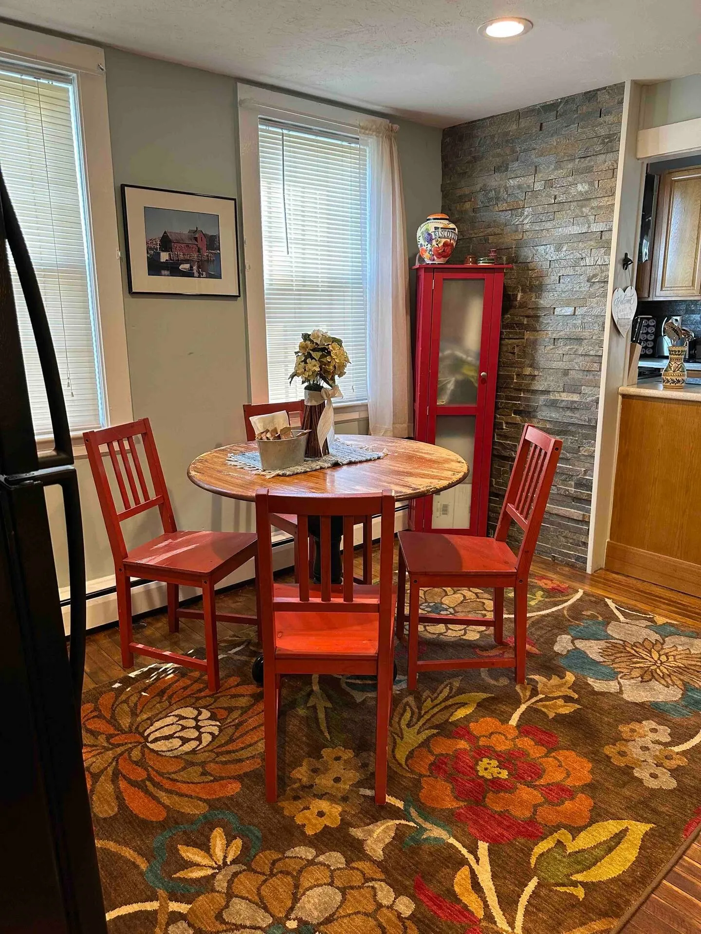 Dining nook with a round wood-topped pedestal table, red chairs, and a floral area rug beside two windows and a stone accent wall.