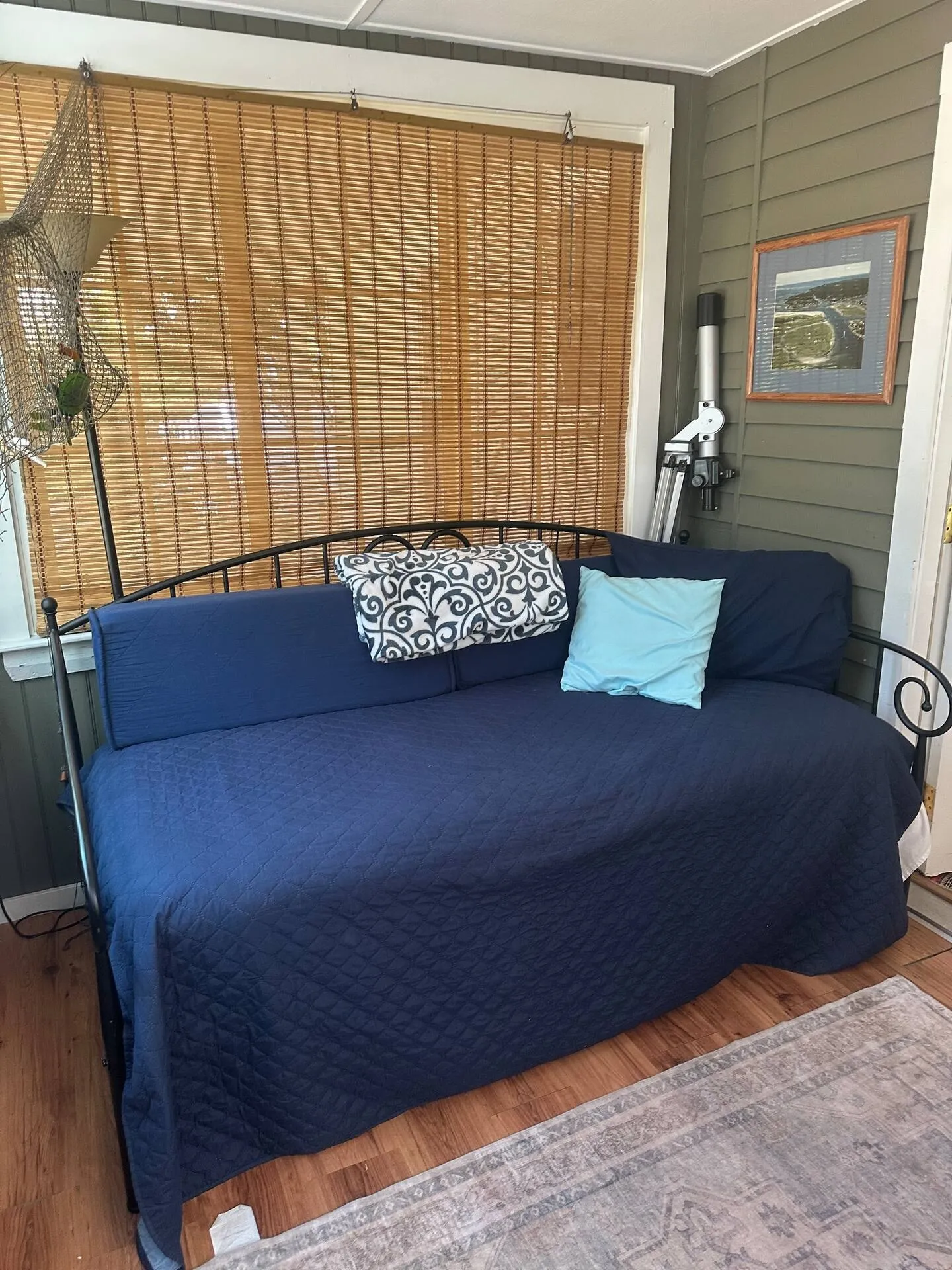 A metal-framed daybed with a navy blue quilted cover and accent pillows situated in a sunroom next to bamboo-shaded windows.