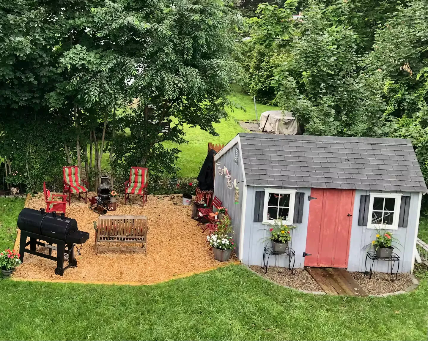 An aerial view of a backyard featuring a small blue shed with a red door next to a wood-chipped seating area with chairs, a chiminea, and a barbecue grill.
