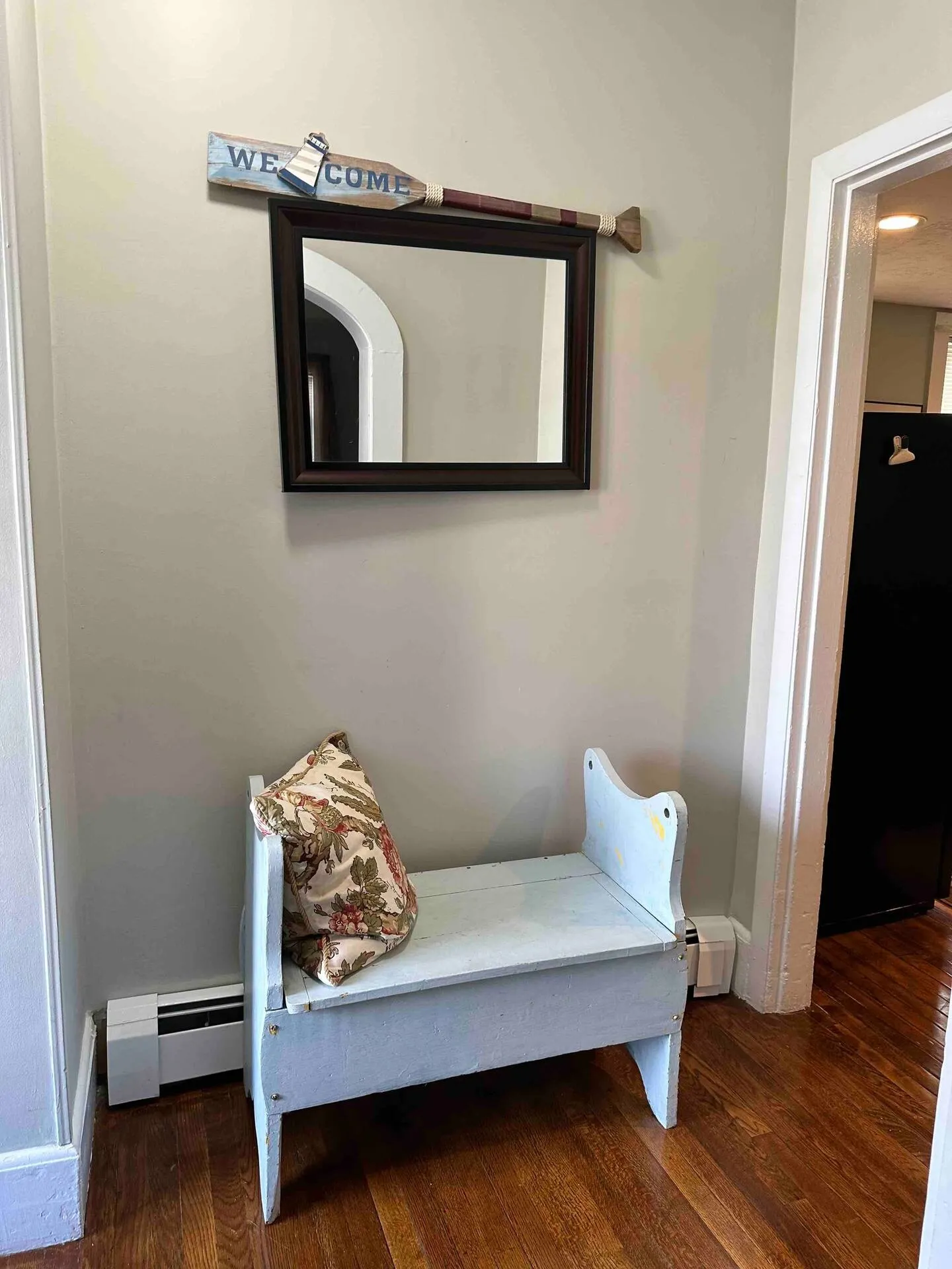Entryway nook with a light blue wooden bench, a patterned throw pillow, a wall mirror, and an oar-shaped Welcome sign.