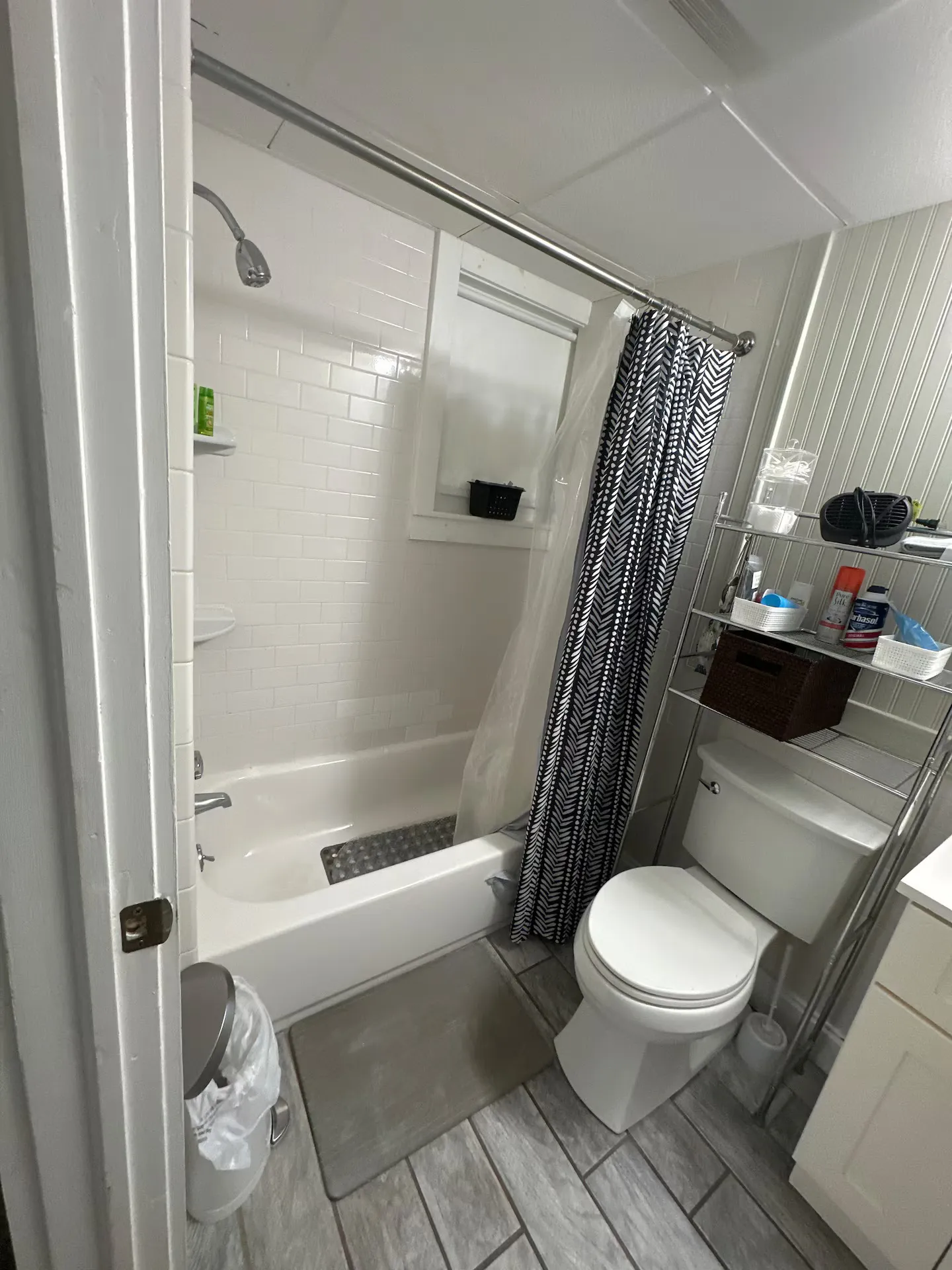 Bathroom with white tiled shower and tub combo, a toilet, wire over-the-toilet shelving unit, and patterned shower curtain.