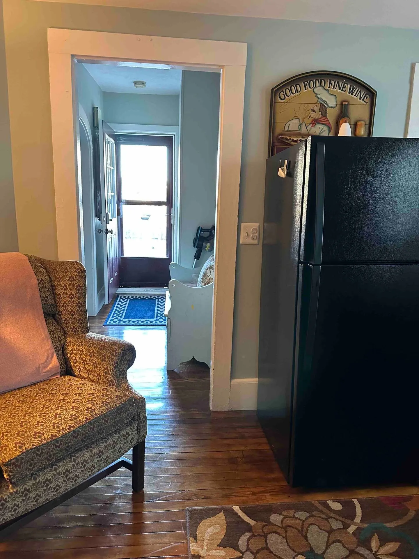 View through a doorway leading into a hallway and front door, showing an armchair, refrigerator, and hardwood flooring.