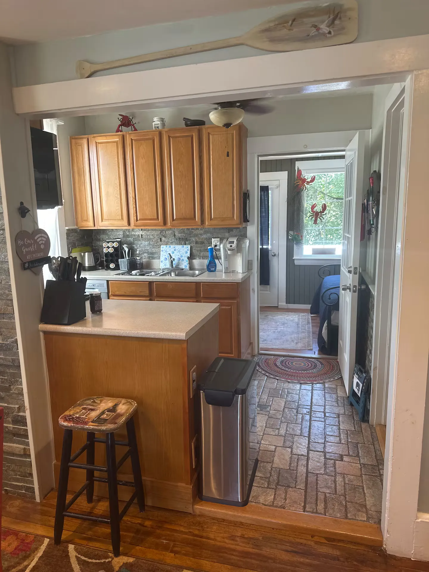 A view through a doorway into a kitchen with wood cabinets, countertop appliances, and a tiled floor leading to an adjacent room.