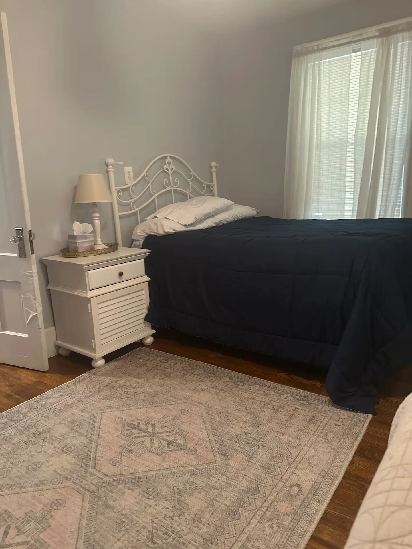 Bedroom with a white metal headboard, dark blue comforter, white nightstand with lamp, and an area rug over wood flooring.