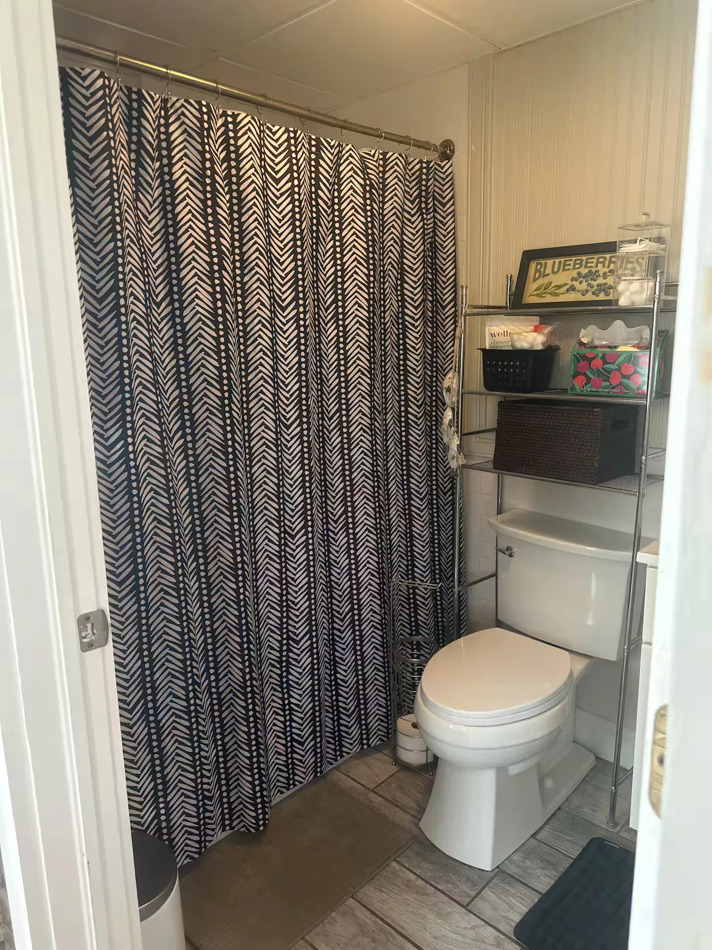 Bathroom showing a toilet beneath a metal shelving unit next to a shower with a patterned dark curtain.