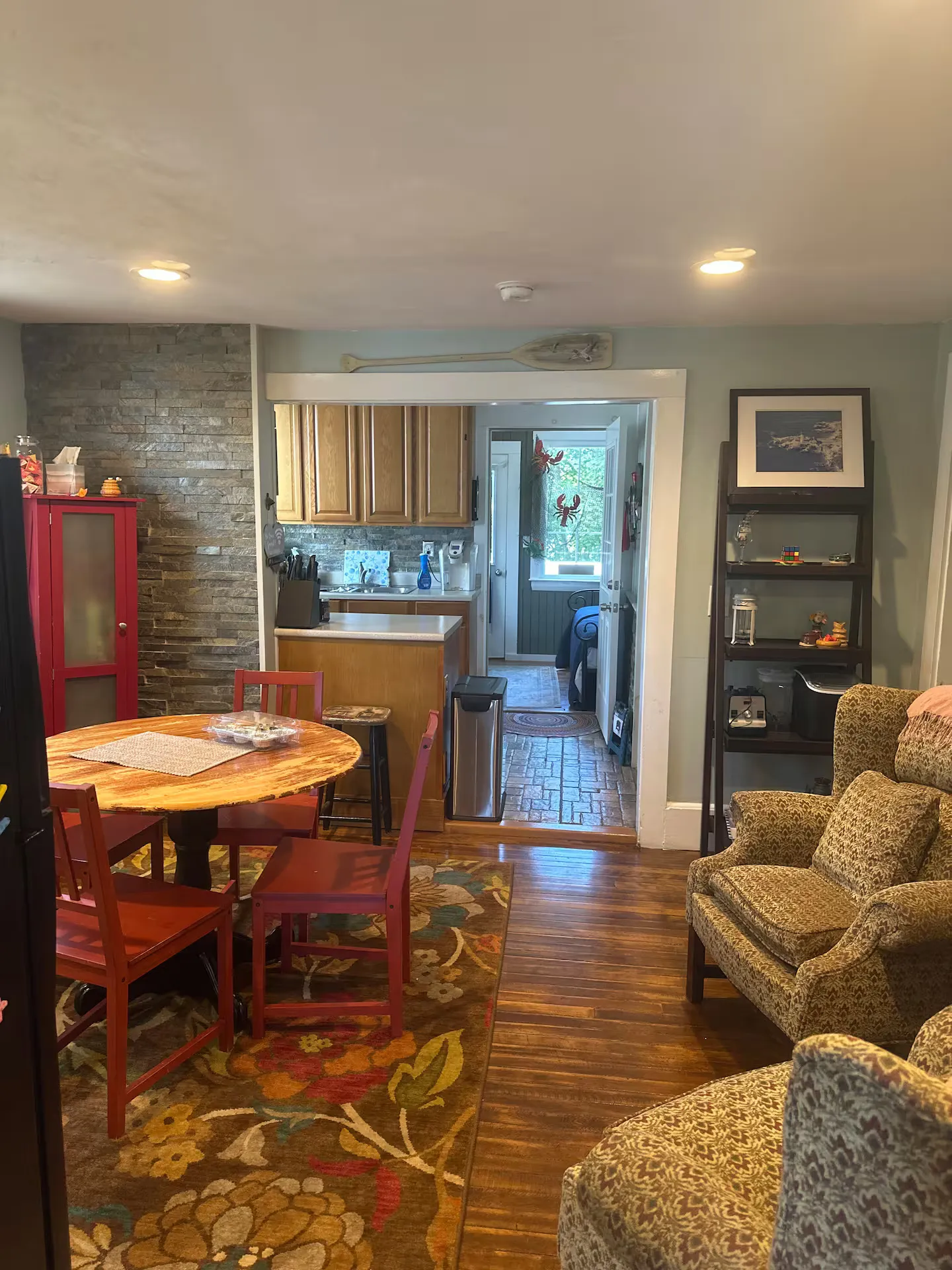 Dining and living area with hardwood floors, a round wooden dining table, patterned armchairs, and a view into the kitchen.