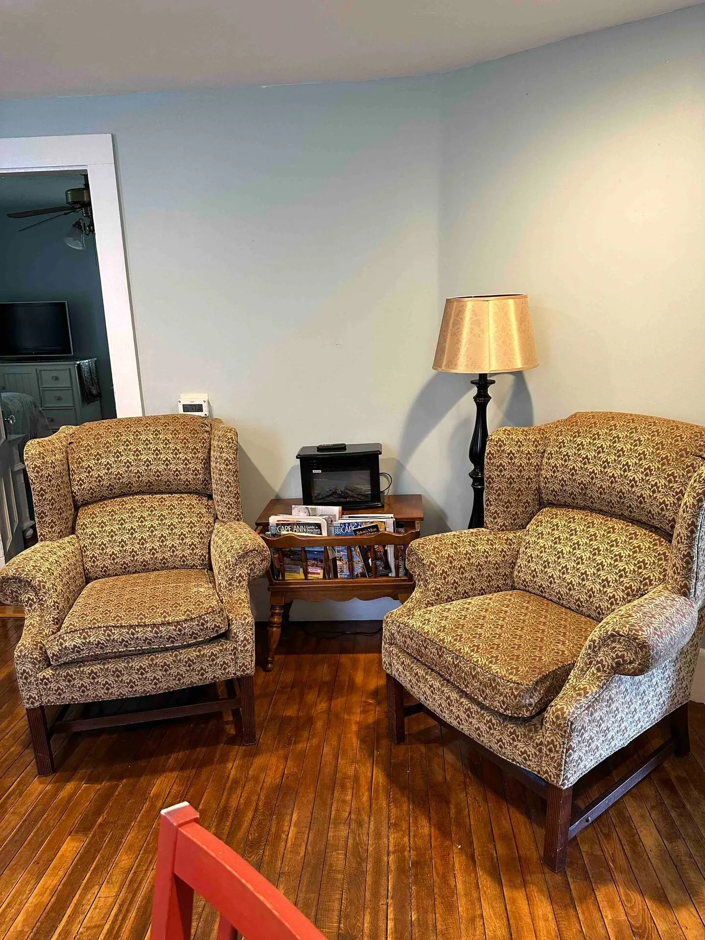 Two patterned wingback armchairs flank a small wooden side table with a standing floor lamp in a room with wood flooring.