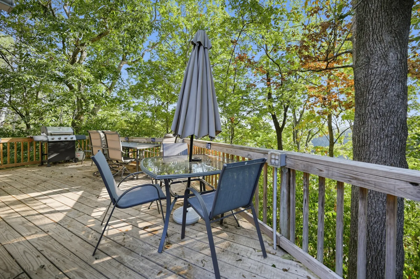 Outdoor wooden deck with outdoor dining tables, chairs, a patio umbrella, and a barbecue grill surrounded by lush green trees.