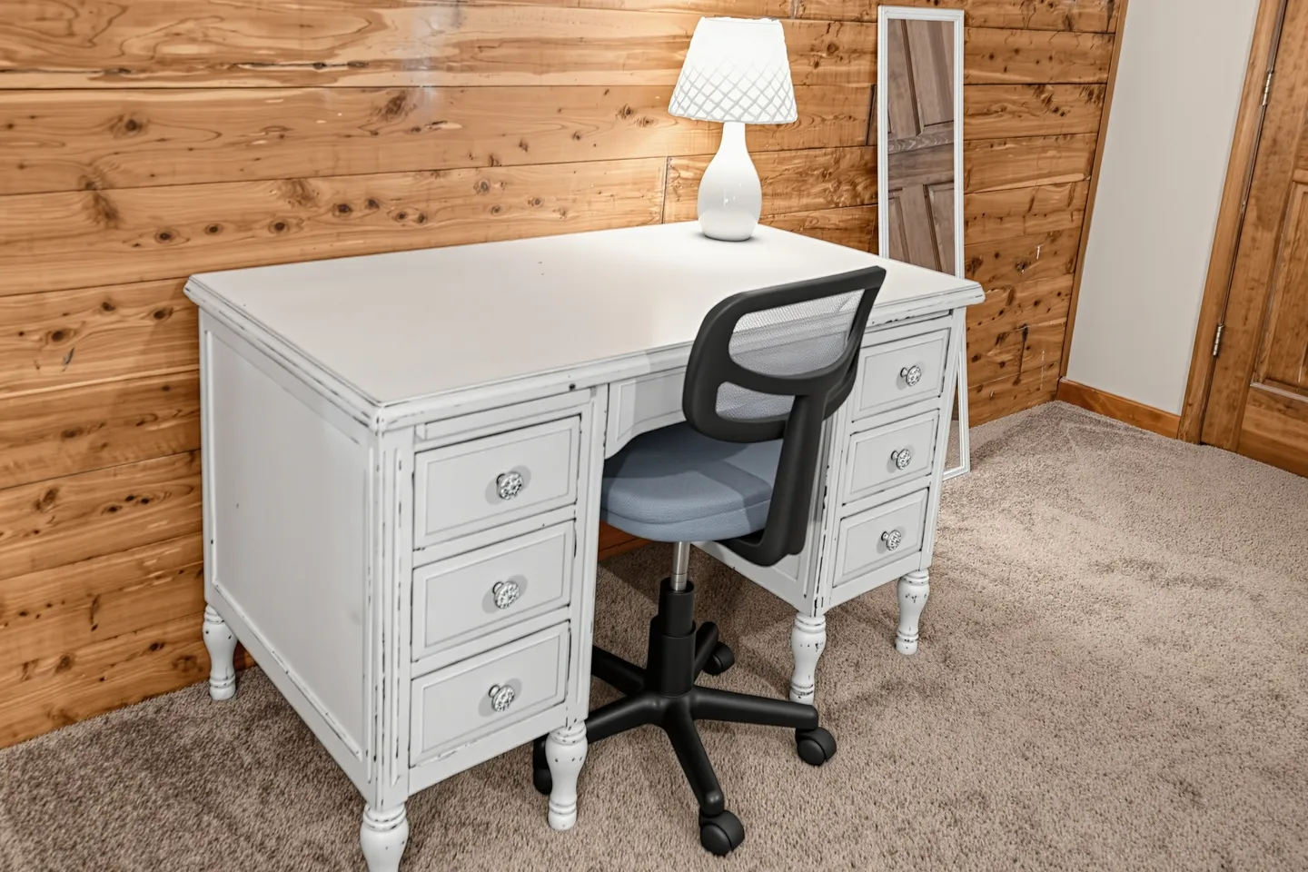 A distressed white desk with drawers, an office chair, a table lamp, and a standing mirror against a wood-paneled wall on carpet.