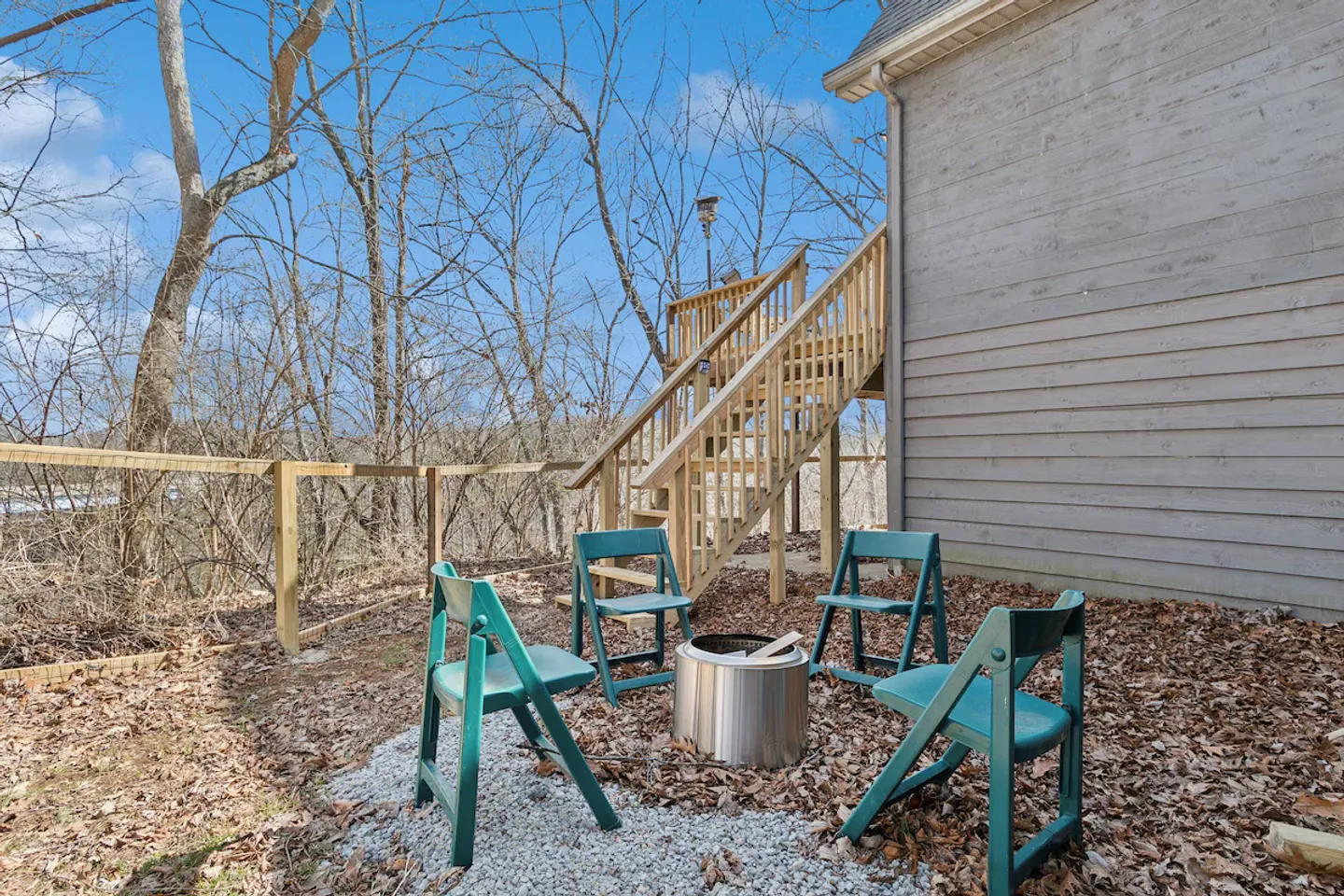 Outdoor fire pit area with four teal chairs and a gravel patch next to a wooden staircase and exterior wall of the house.