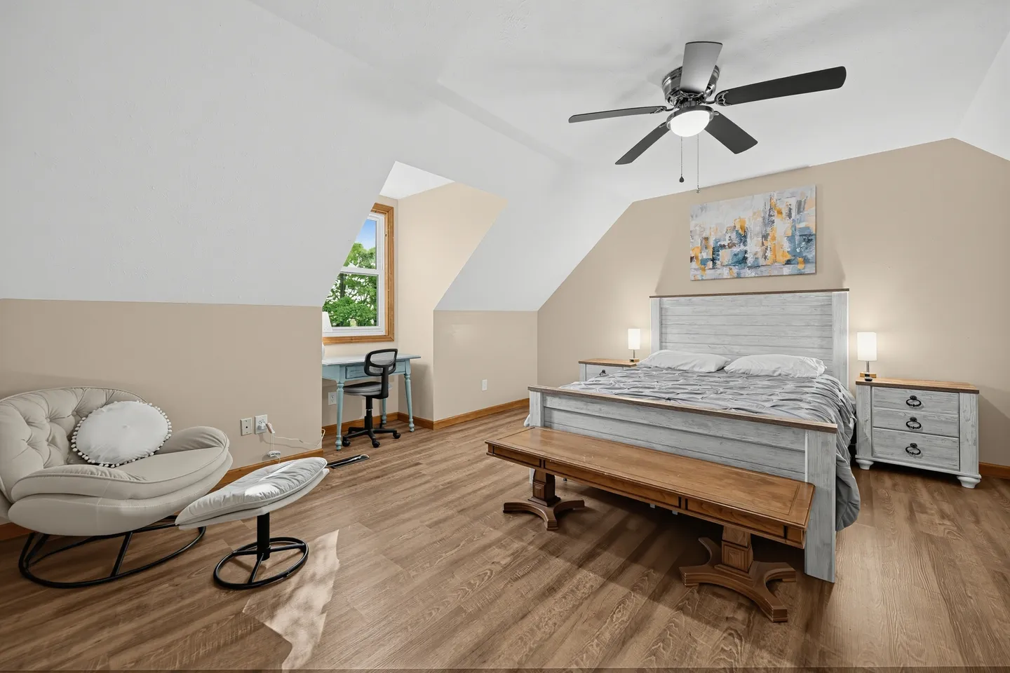 Spacious bedroom with sloped ceilings, a light wooden bed frame, a wooden bench, and a desk nook by a window.
