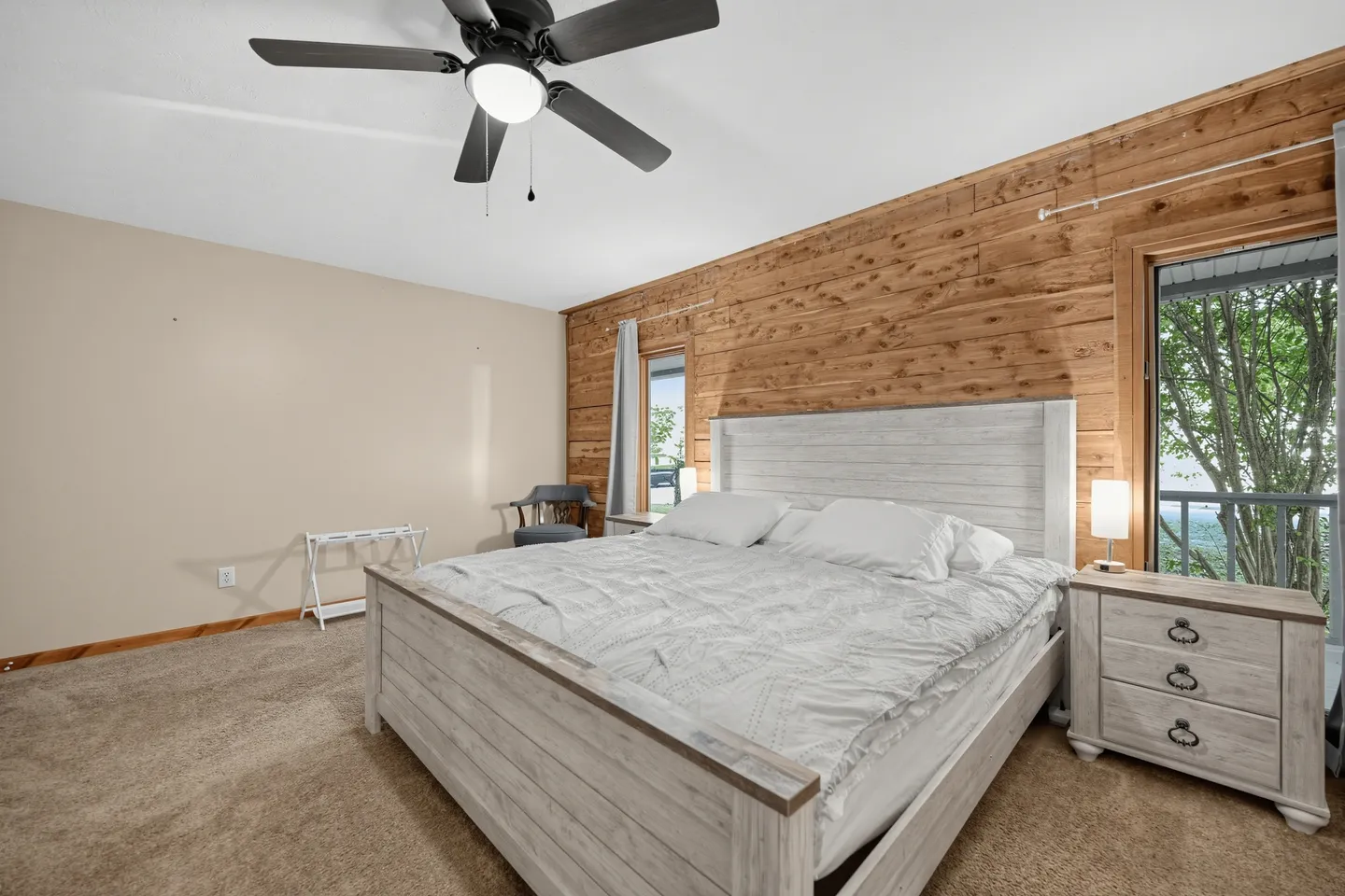 Bedroom with a white wooden bed frame, wood-paneled accent wall, bedside table with lamp, ceiling fan, and carpeted floor.