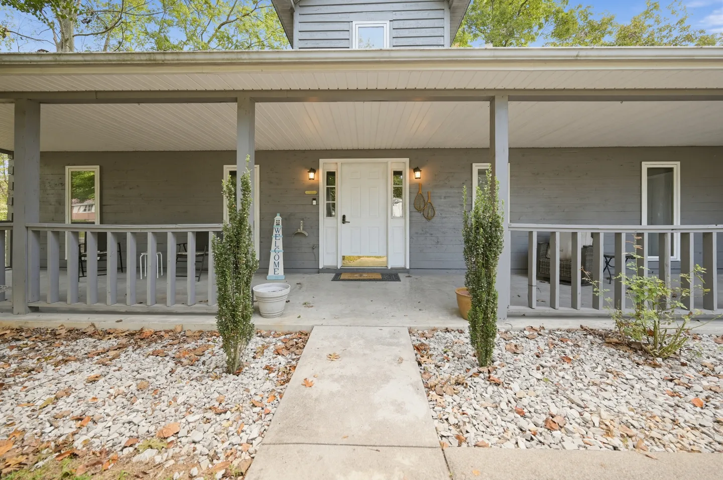 Front porch of a gray house with a white front door, railing, and a concrete walkway flanked by small shrubs and gravel landscaping.