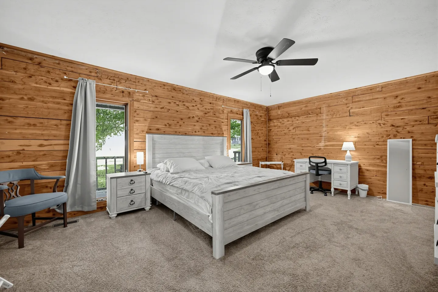 Bedroom with wood-paneled walls, carpet, a king-size bed, a bedside table, an armchair, and a white desk with an office chair.