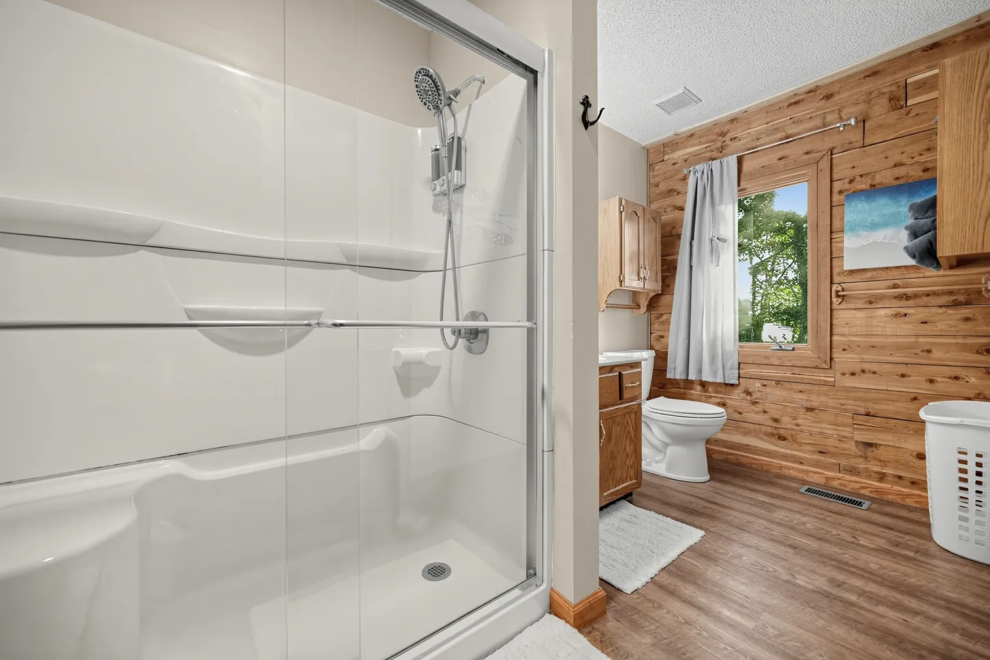 Bathroom with a glass-enclosed shower stall on the left, wood-paneled accent wall, window, vanity, and toilet.