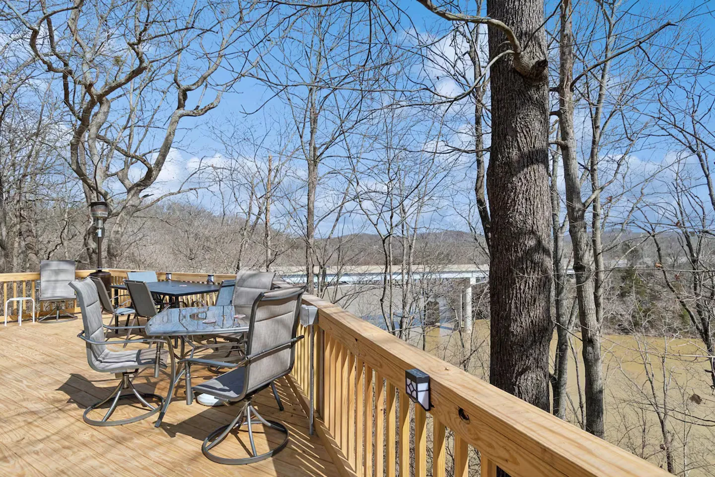 Elevated wooden deck with outdoor patio tables, swivel chairs, and views of a river and bridge through bare trees.
