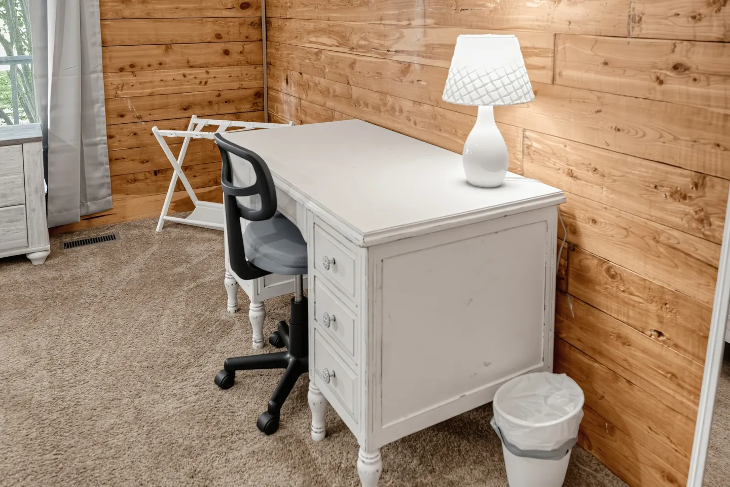 A white painted desk with drawers, an office chair, and a table lamp against a wood-paneled wall.