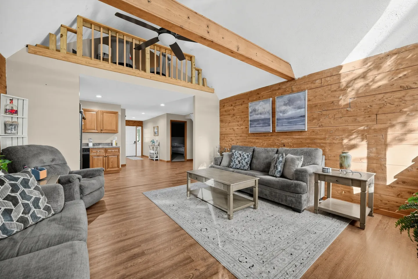 Open-concept living room with a wood-plank accent wall, loft railing, exposed wooden ceiling beam, and gray seating.
