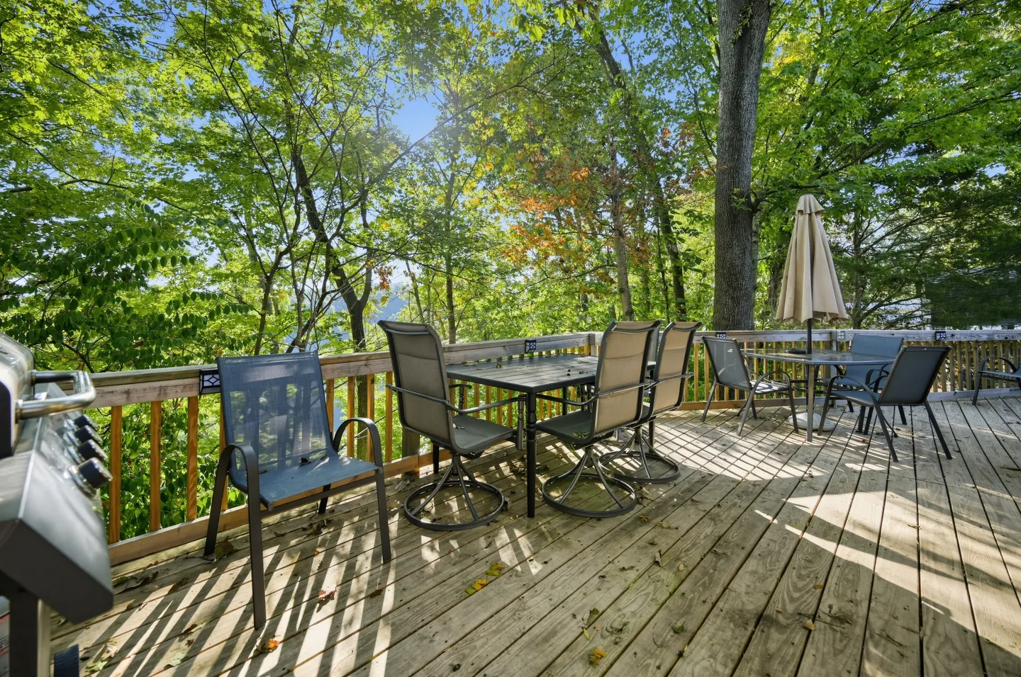 Outdoor wooden deck with patio dining tables, chairs, a barbecue grill, and surrounding lush green trees.