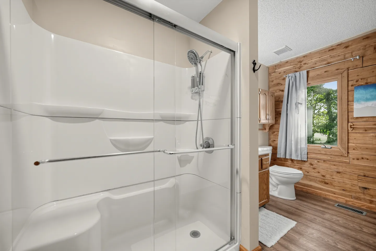 Bathroom with a sliding glass door walk-in shower in the foreground and a toilet and wood-paneled wall in the background.
